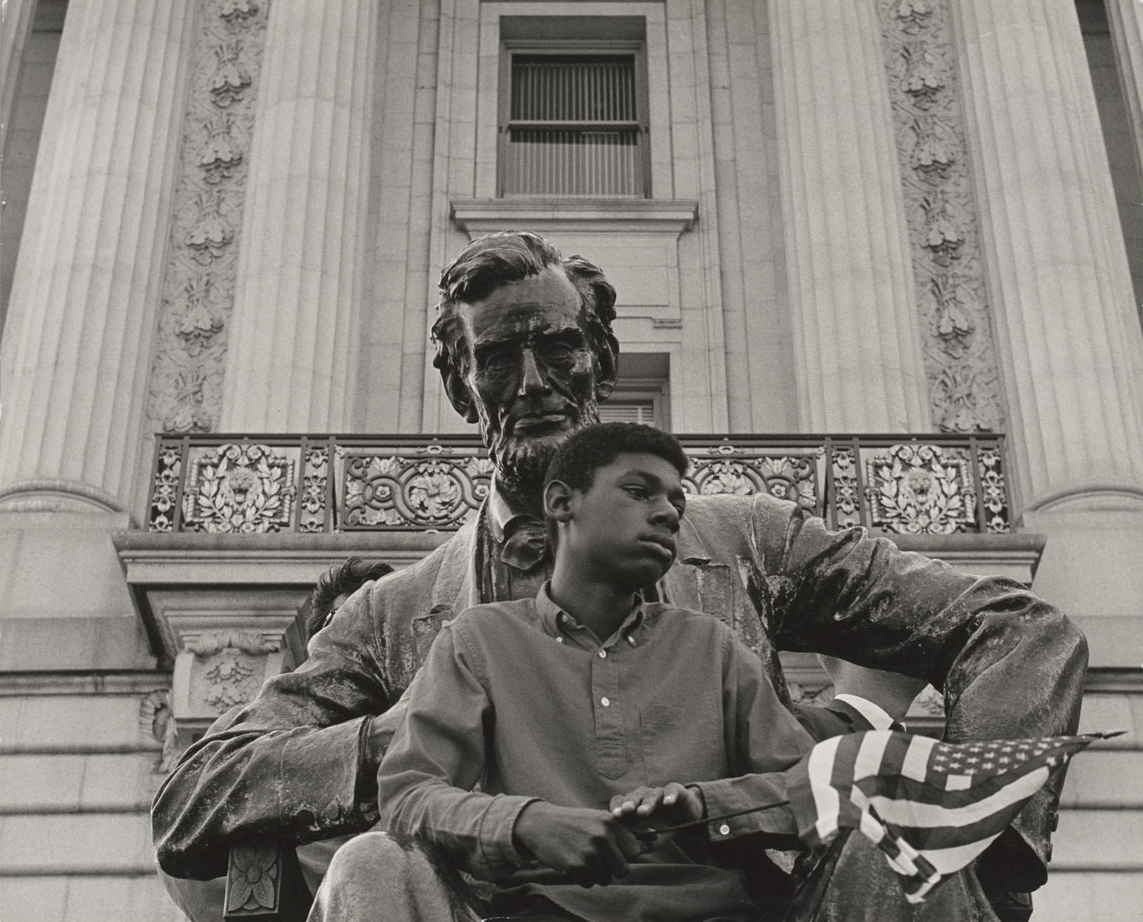 An African-American child holding a flag and sitting on the lap of a statue of Abraham Lincoln